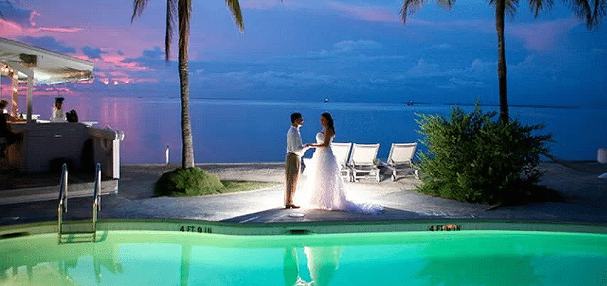 A couple stands near the poolside at Casa Morada with lounge chairs and palm trees in the background during sunset.