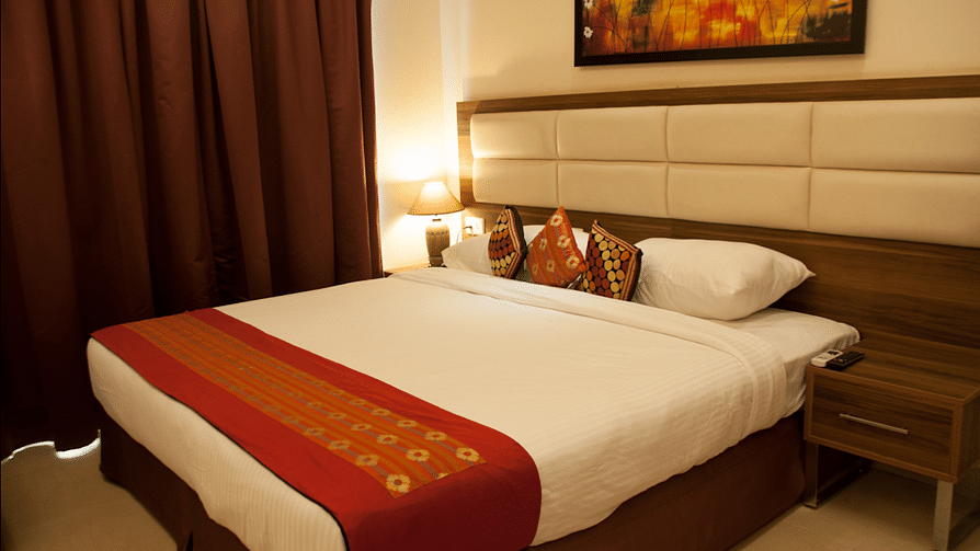 A neatly made hotel room bed with a decorative red runner and pillows, beside a nightstand and drawn curtains | Astra Hotels and Suites Electronic City