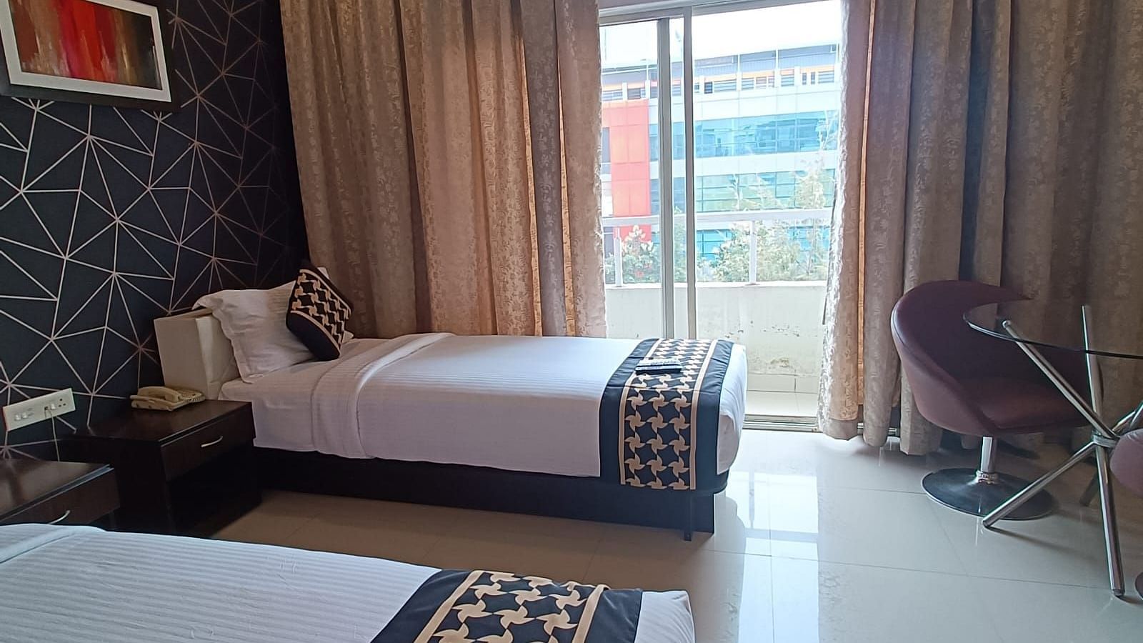 Hotel room designed for comfort and style, with two beds dressed in matching linens, a bold accent wall adding a touch of contemporary flair, and a huge glass window with an outdoor view, along with comfortable seating for relaxation - Astra Hotels & Suite
