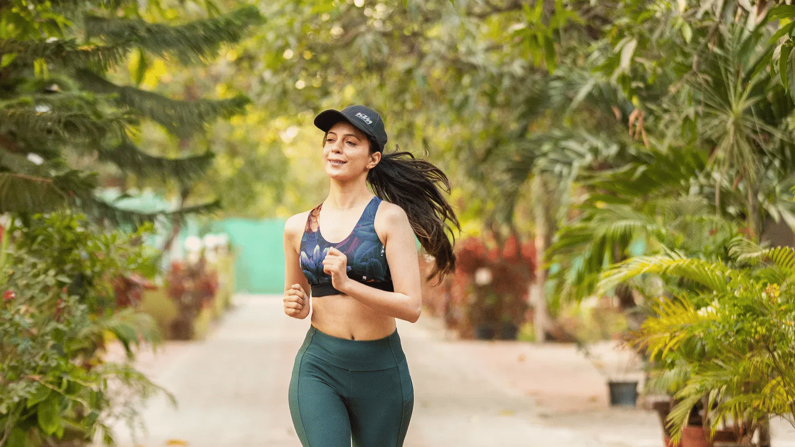 A woman wearing active wear jogging on a clear pathway lined completely by plants and trees -Daksh Eden Greenz, Sasan Gir