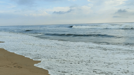White sea foam and gentle ocean waves crashing onto a sandy beach under a light blue sky with scattered clouds.