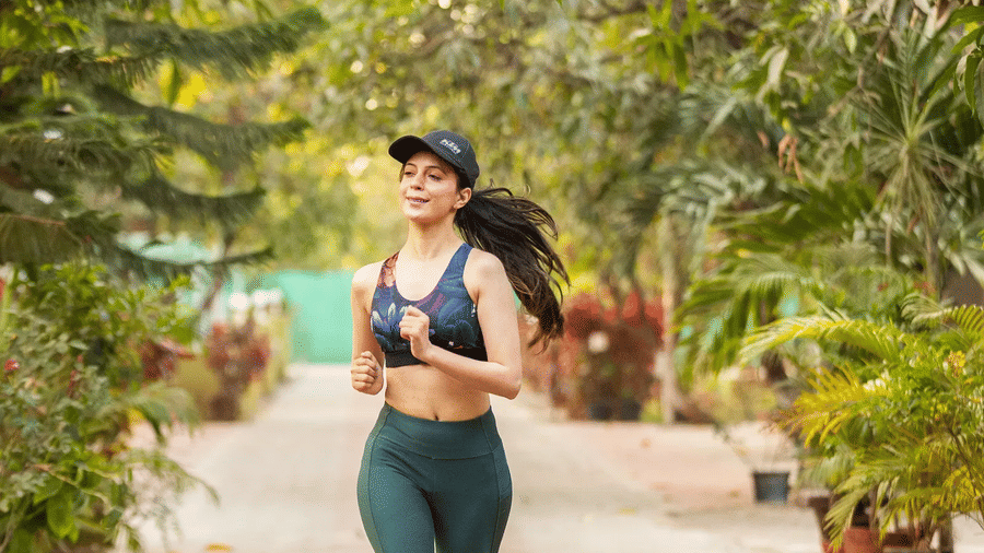 A woman wearing active wear jogging on a clear pathway lined completely by plants and trees -Daksh Eden Greenz, Sasan Gir