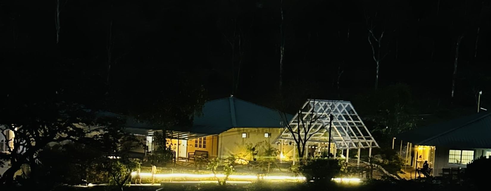 A wide shot of a property at night, illuminated by numerous warm yellow lights outlining pathways with dark trees in the background.