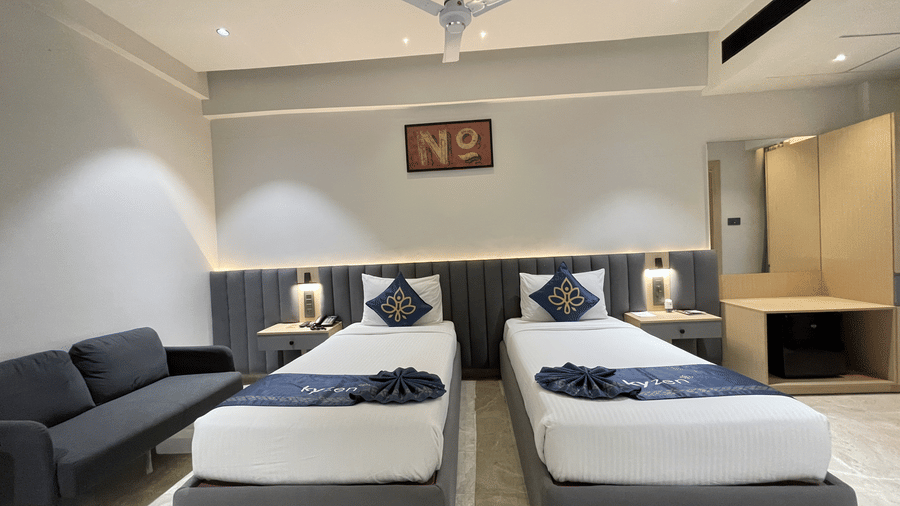A twin bed room at Hotel Kyzen HiTech City featuring two beds with blue accent bedding grey upholstered headboards a grey sofa ceiling fan and framed artwork on the wall.