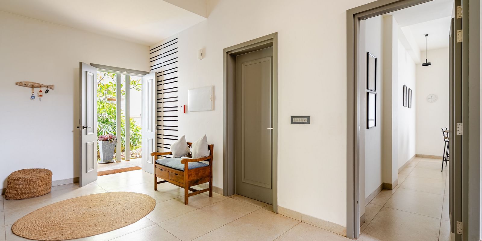 An entryway with an open front door leading to a patio, featuring a small wooden bench with pillows, a round woven rug, and a closed grey door inside - Lindsay Manor