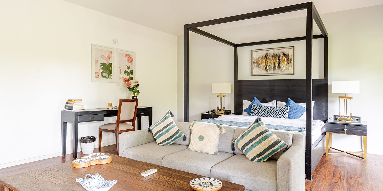 Bright and airy room featuring a modern four-poster bed with accent pillows, a neutral-toned sofa with striped cushions, a wooden coffee table, and a black writing desk with floral décor and framed botanical art - Lindsay Manor