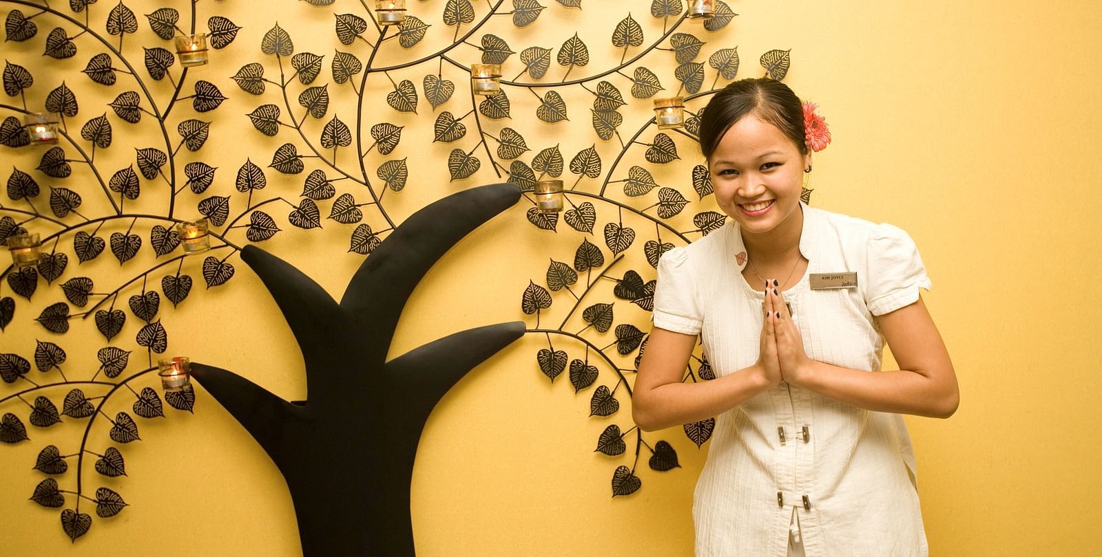  A lady greeting guests in front of a wall which has a tree poster at Grand Chennai By GRT Hotels.