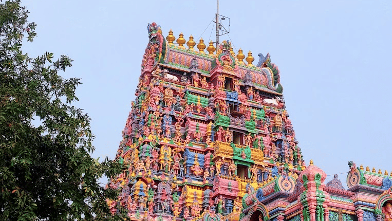 A tall, intricately carved and brightly painted Pillayarpatti Temple's gopuram with colourful statues and decorative details rising against a clear blue sky, partially framed by tree foliage near Chidambara Vilas.