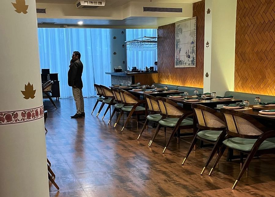 Modern restaurant interior with long, neatly arranged tables, decorative pillars featuring lotus designs, and a glimpse of a person through a window in the background - Astra Hotels & Suites, Spice Garden Layout