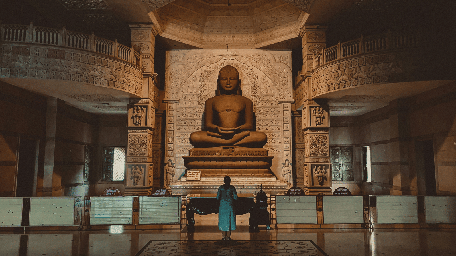 An inside view of a jain temple with a deity in the background and a person praying in front of it.