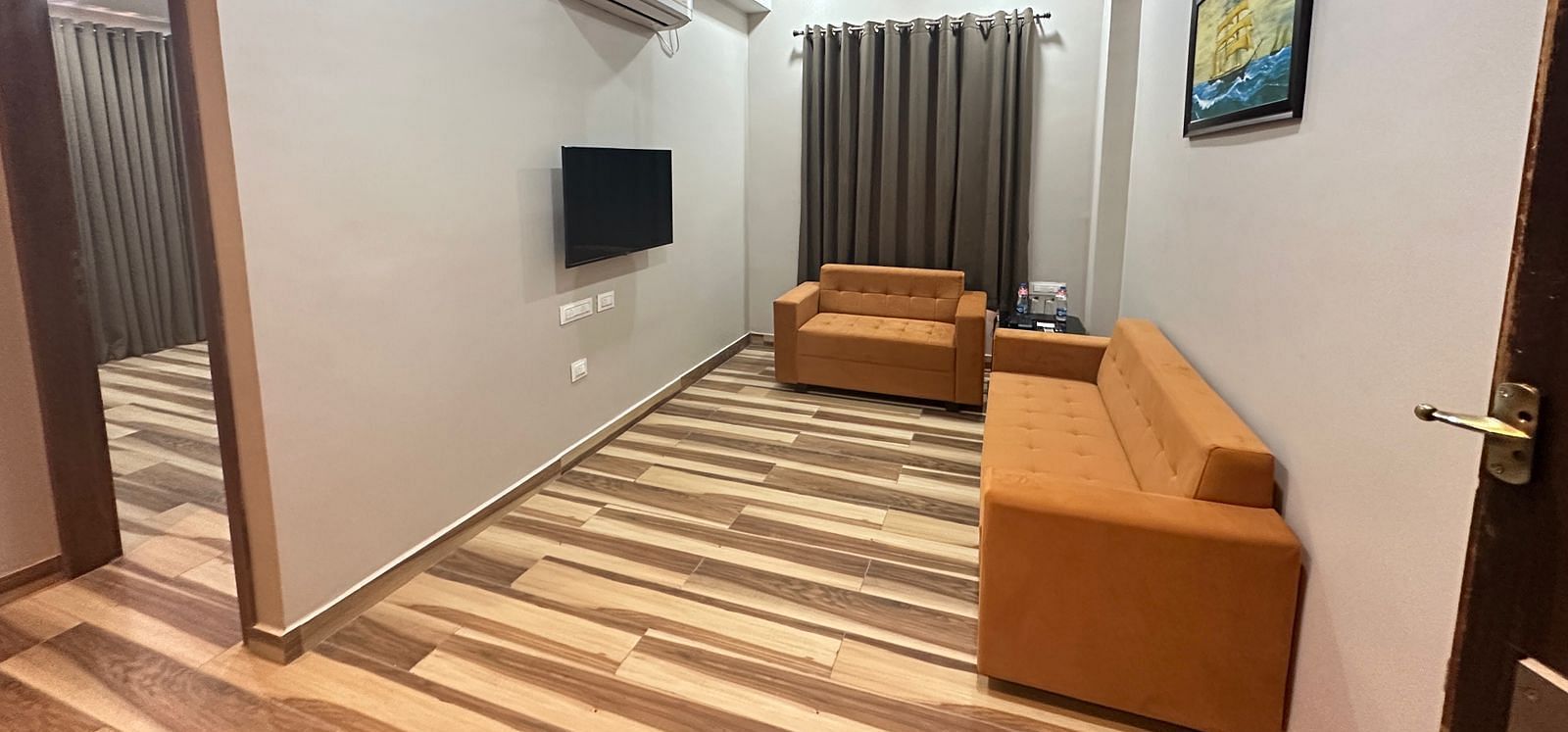A room features a double bed, a brown sofa, a television, and wooden flooring - Kankidham Lords Inn