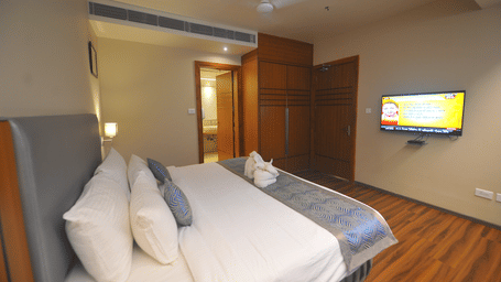 Suite bedroom with a bed dressed in white and blue linen, a grey upholstered headboard, a wall-mounted television and dark wooden flooring.