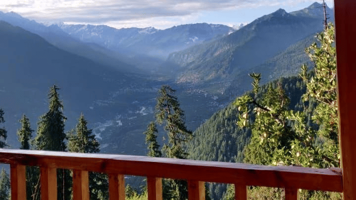 A beautiful scenery from the wooden balcony of Amara Upepo - The Sky Village, Manali, looking out at a mountain valley with layers of forested slopes under a bright, cloudy sky.
