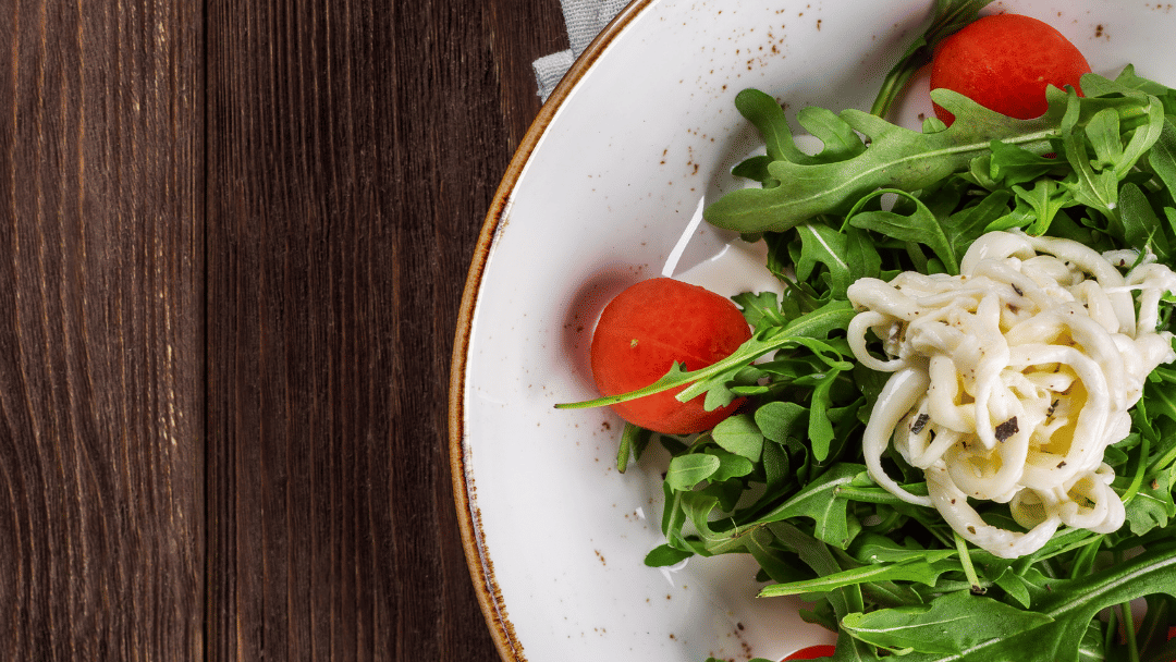 Fresh arugula salad with cherry tomatoes and soft cheese served on a white plate over a rustic wooden table.