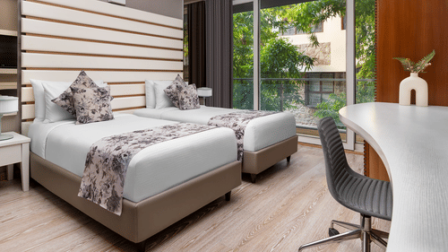  Contemporary room at The Atara, Gurugram featuring two single beds, a white desk, and a balcony view. The room has light wood floors and horizontal wall decor