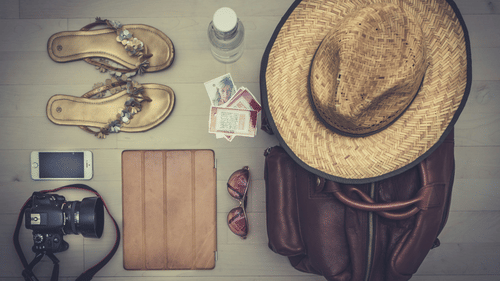 A collection of travel items, including a straw hat, sandals, a bag, and a camera, laid out on a wooden surface.