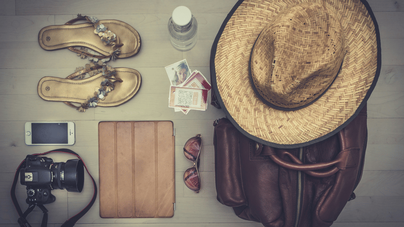 A collection of travel items, including a straw hat, sandals, a bag, and a camera, laid out on a wooden surface.