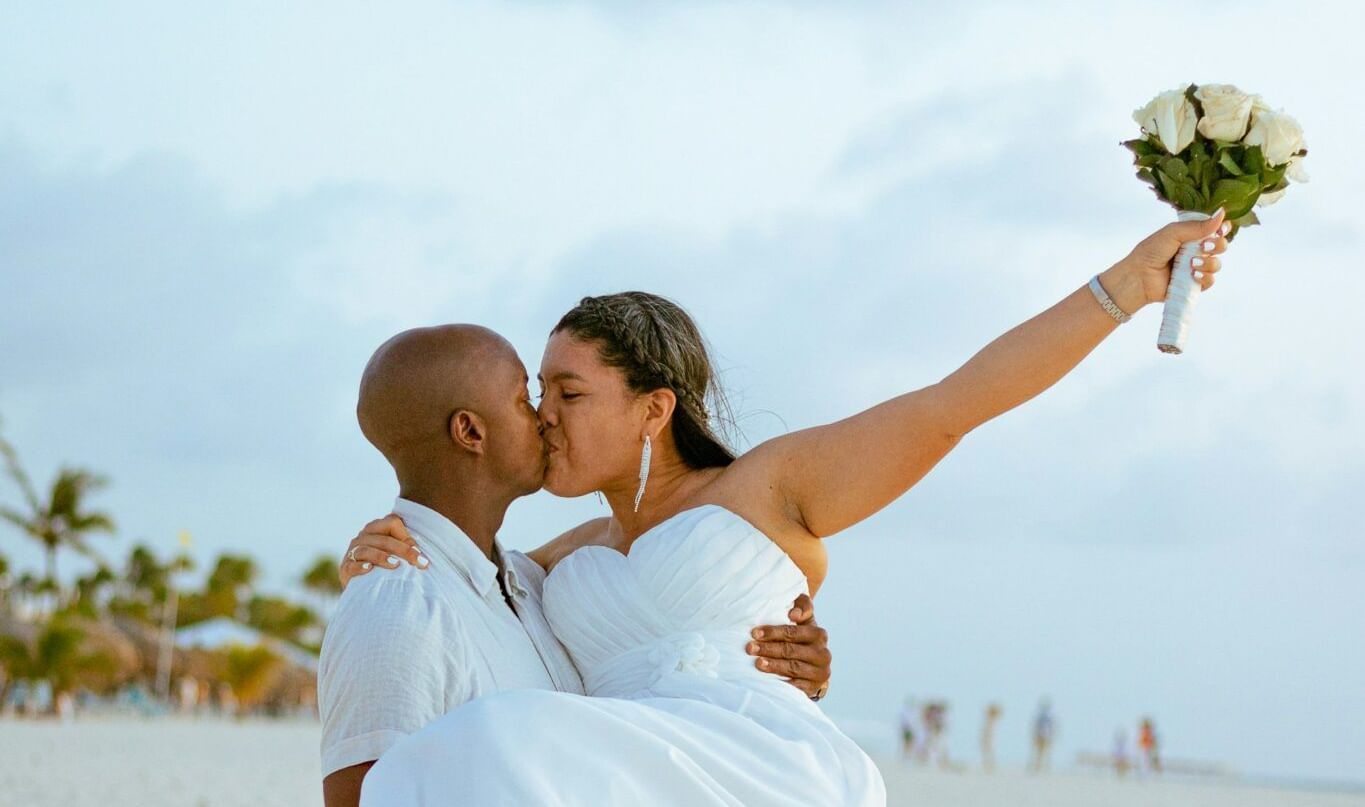 A couple celebrating on a beach, with the groom carrying the bride in his arms against a backdrop of the ocean.