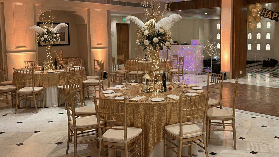 A banquet hall with round tables, gold-toned chairs, floral centerpieces, and a decorated stage backdrop at Clarks Avadh, Lucknow.