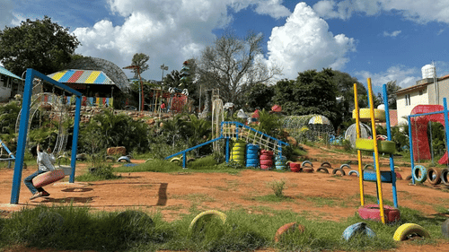 A colourful outdoor playground with swings, climbing structures, and painted tires under a bright blue sky with fluffy clouds. A child sits on a tire swing. Grass and trees surround the area.