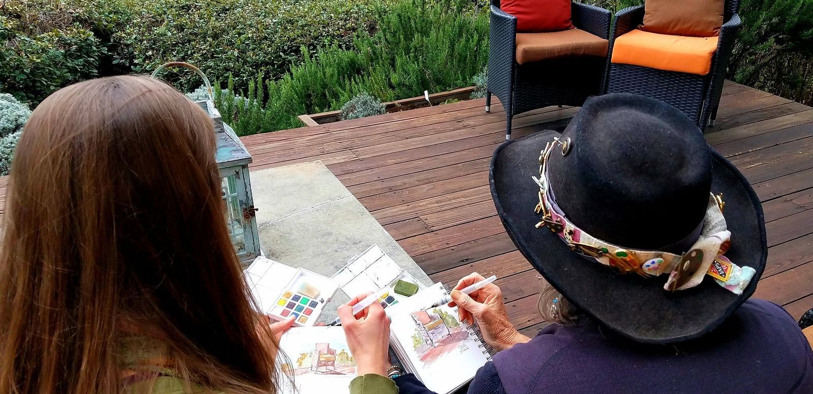 Two women sitting outdoors with a scenic ocean view, engaged in painting or sketching, with one wearing a black hat adorned with flowers.