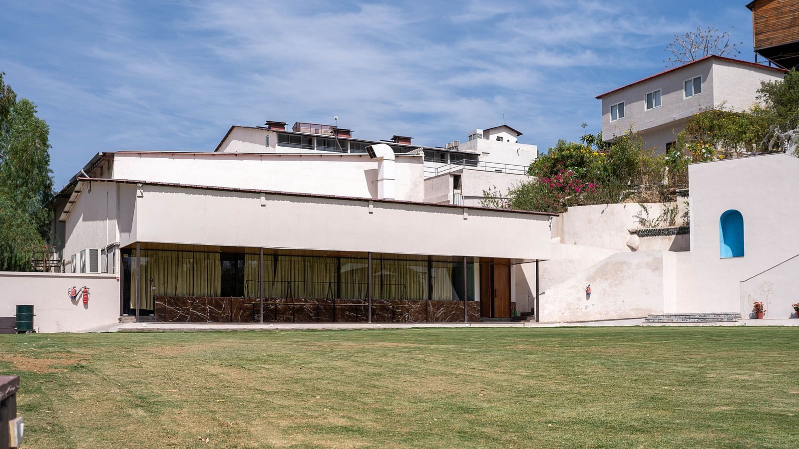 A facade of the white building of Bamboo Saa Resort & Spa, Udaipur with a flat roofline and a blue door set against a blue sky with white clouds.