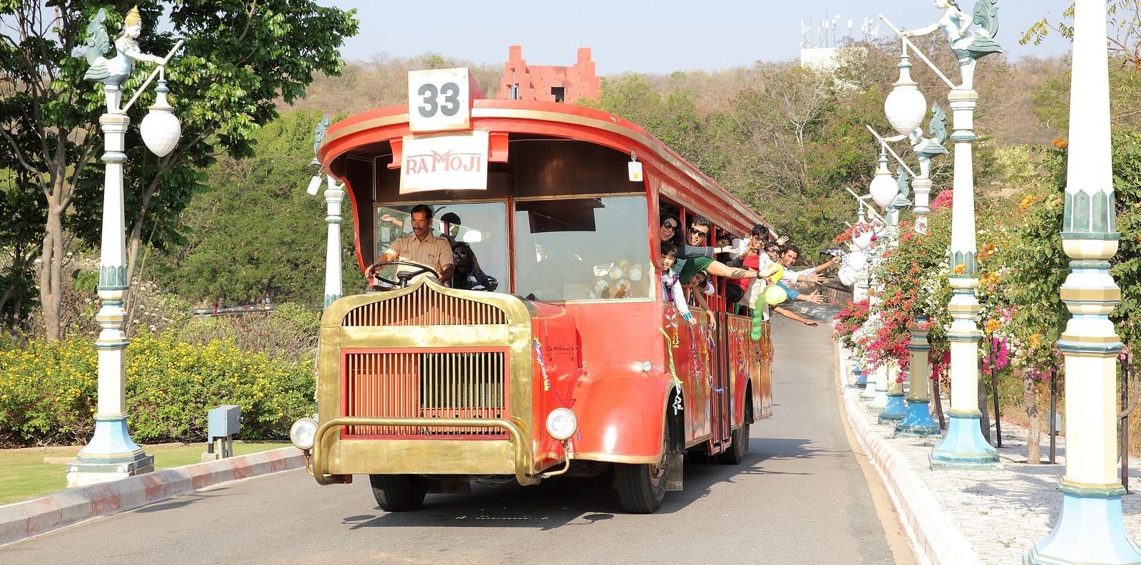 A red vintage-style trolley bus carrying visitors through a well-manicured amusement park pathway, offering a charming and nostalgic ride at Ramoji Film City Hotels in Hyderabad 