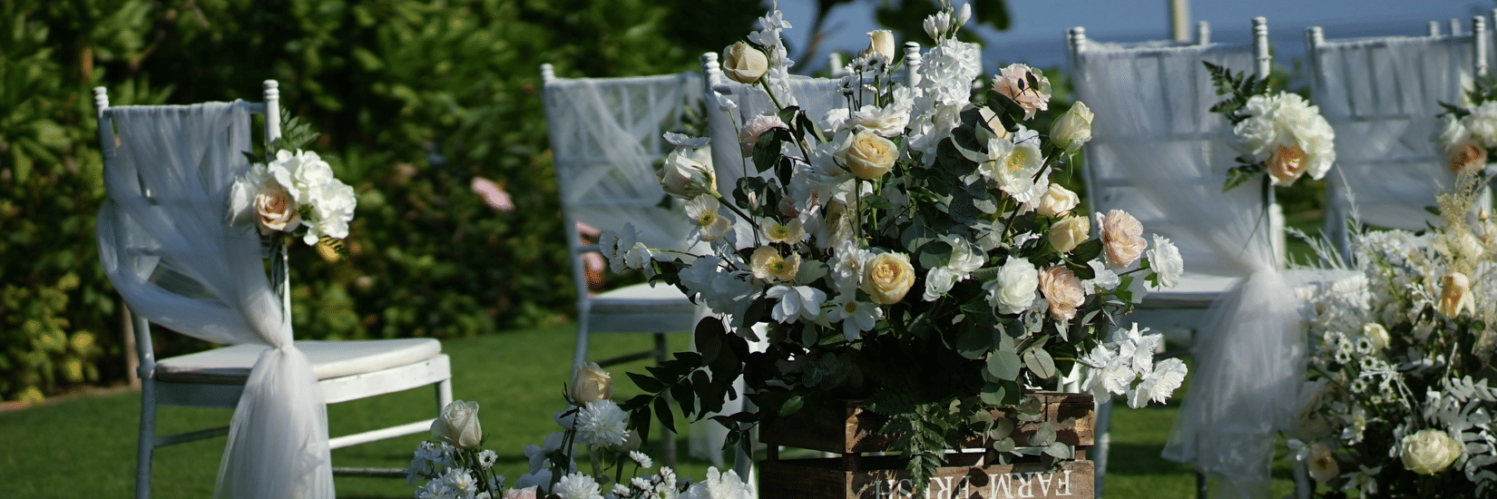Rose petals cover green grass in a sunny outdoor wedding setting with chairs draped in fabric and large floral arrangements in wooden crates.
