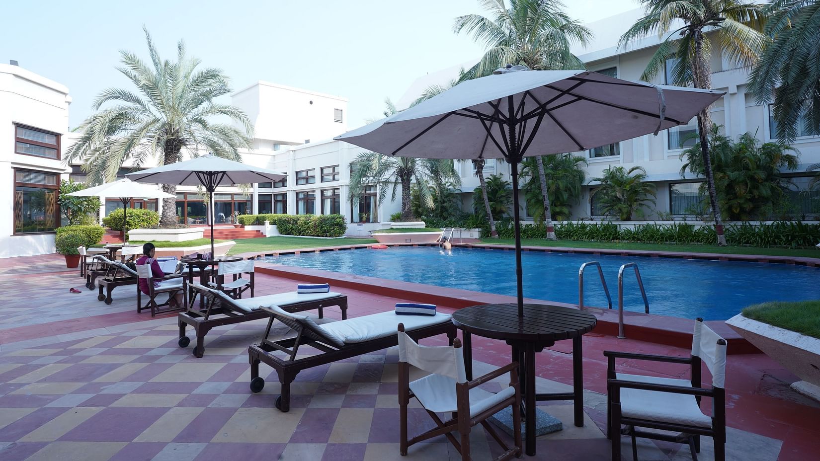 A swimming pool surrounded by palm trees and sun loungers, with a hotel building visible in the background -The Ummed Ahmedabad