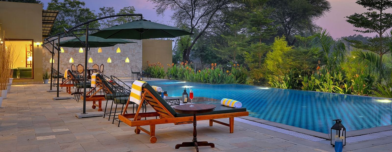 A poolside scene at dusk features lounge chairs with striped towels, small tables with wine glasses, and umbrellas beside a blue-tiled swimming pool surrounded by lush greenery and illuminated by string lights - Singhvilas, Ranthambore