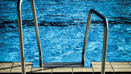 Close-up of metal steps leading into a shimmering blue swimming pool from a paved edge.