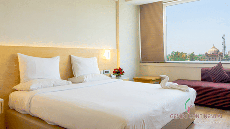 Hotel room with a bed dressed in white linen, a maroon sofa placed nearby, a large window with a city view and light-toned interiors all around.