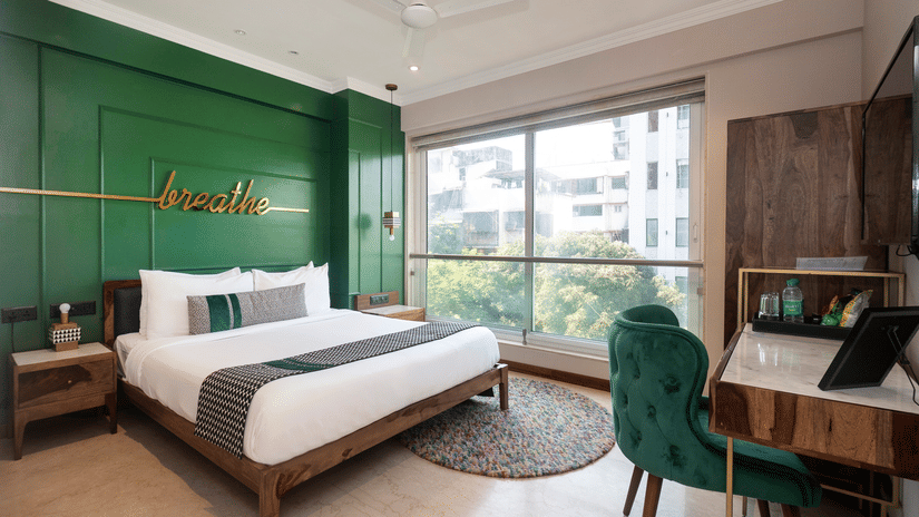 A bedroom at Theory9 - Premium Service Apartments, Bandra, featuring a large bed with white linens and a green accent wall with the word 'breathe' in gold, a wooden nightstand, a desk with a green velvet chair, and a large window overlooking trees.