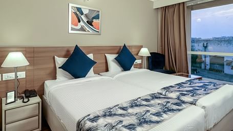 Premium Studio at Starlit Suites Newtown, Kolkata features two single beds with blue and white patterned throws and cushions, bedside tables with lamps, and a large window.