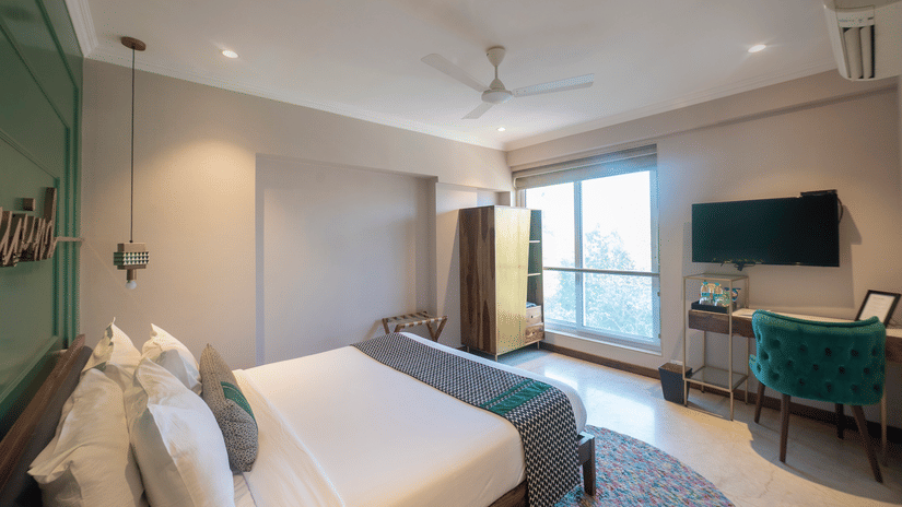 A bedroom with a neatly made bed, pendant lighting, ceiling fan, wardrobe, work desk with chair, wall-mounted TV, and a large window letting in natural light - Theory9 - Premium Service Apartments, Bandra