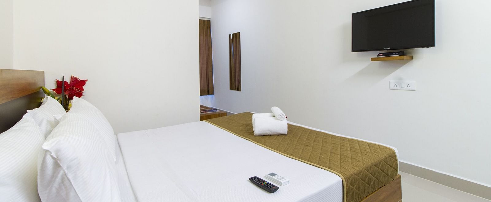 A room showing a double bed with white bedding and a television mounted on the wall at Sanctum Suites Whitefield, Bangalore.
