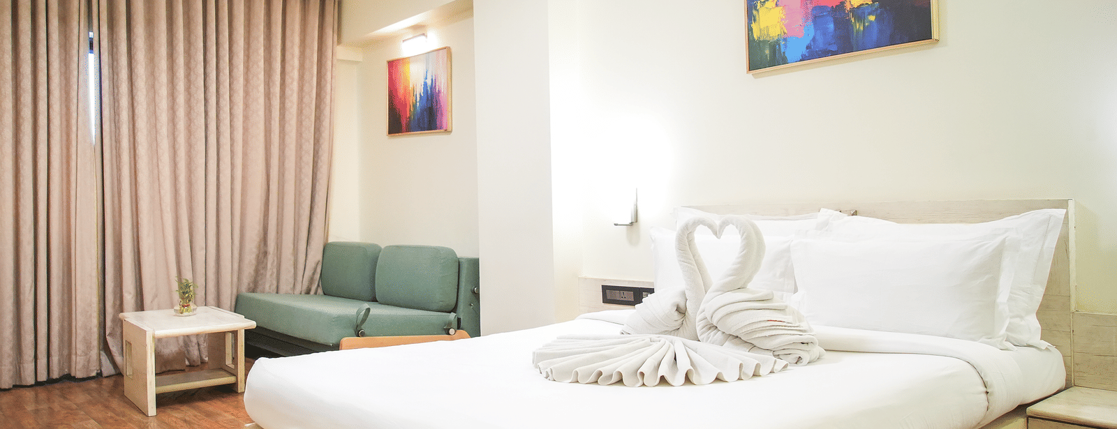 A room with a king size bed, a sofa and a chair, wall arts and light brown curtains at Infiniti Hotel, Indore.