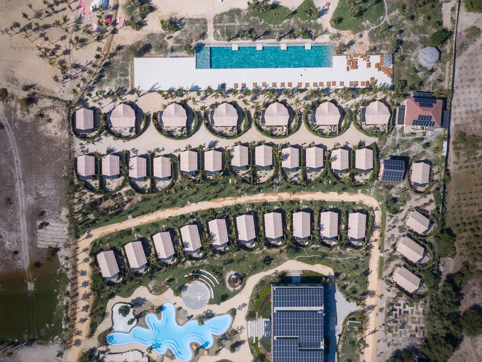 Aerial view of WoCAL Retreat Ke Ga featuring numerous circular bungalows, multiple swimming pools, and manicured green spaces nestled in a dry, coastal landscape.