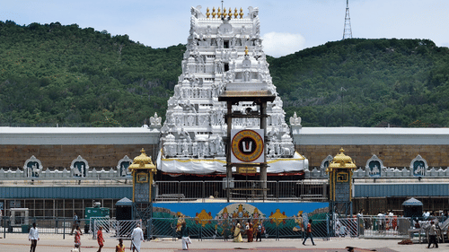 A wide shot of a white, multi-tiered Tirumala Venkateswara Temple tower with golden accents, situated in a courtyard with green hills and a clear sky in the background.