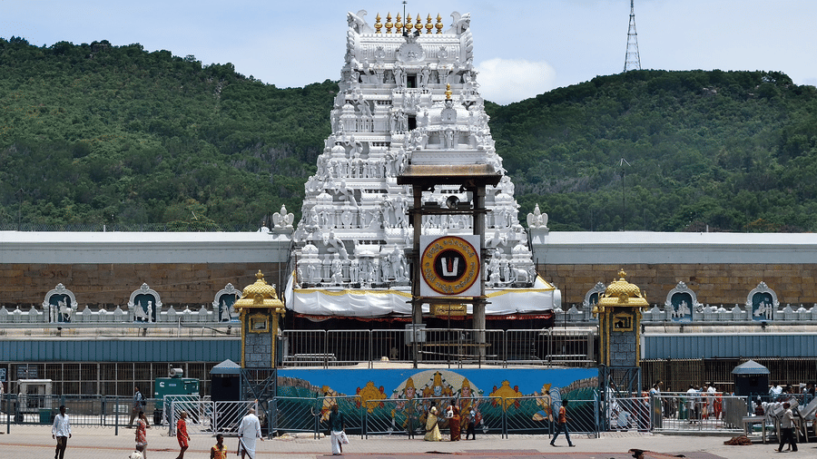 A wide shot of a white, multi-tiered Tirumala Venkateswara Temple tower with golden accents, situated in a courtyard with green hills and a clear sky in the background.