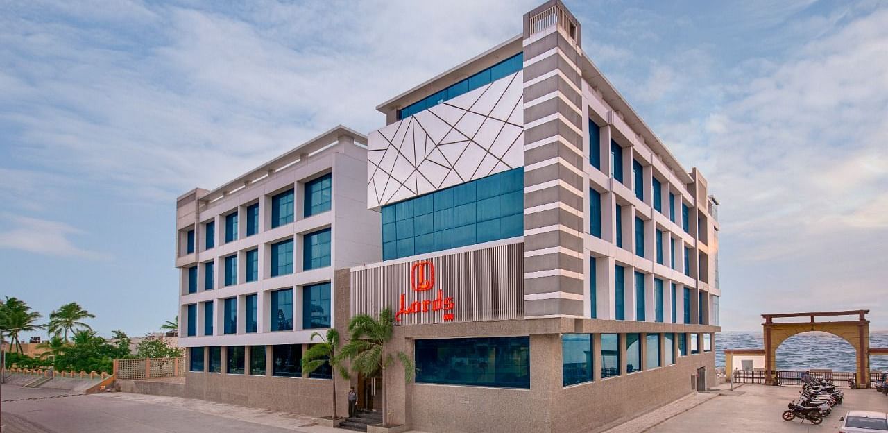 A light-colored hotel with a distinctive architectural style is located near the coast, with gentle waves lapping the shore under a clear sky.