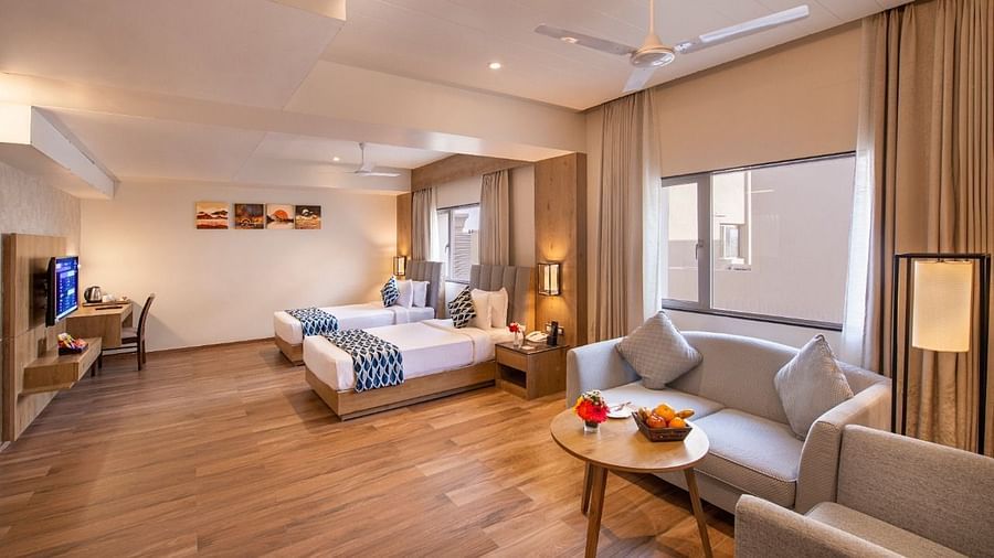 A spacious hotel suite featuring two single beds with blue and white accents, a comfortable seating area with a sofa and armchairs around a small table with fruit, a work desk with a chair, and a television, all on a wooden floor at Lords Inn Motikhavdi