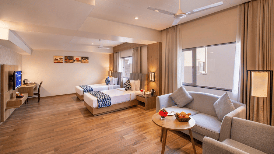 A spacious hotel suite featuring two single beds with blue and white accents, a comfortable seating area with a sofa and armchairs around a small table with fruits in Lords Inn Jamnagar