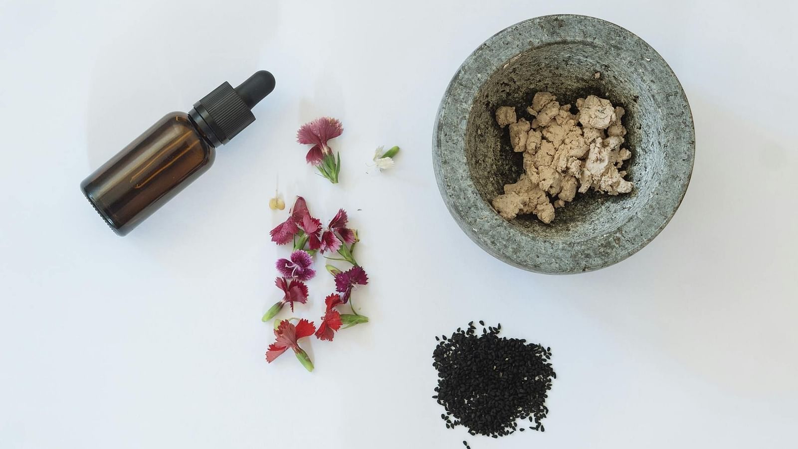 A glass vial, flowers, herbs and a mortar aesthetically placed on a white surface.