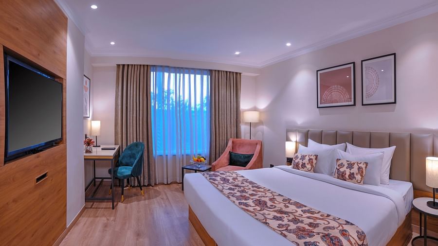 A room with clean bed, a TV, wall paintings, sofa chairs and curtains - Royale Sarovar Portico, Agra