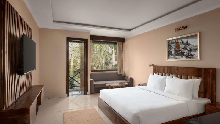 A Bay Room at Corbett Adventure Resort, Ramnagar, featuring a spacious bedroom with a king size bed, comfortable seating area, television, and large windows opening to lush outdoor views.