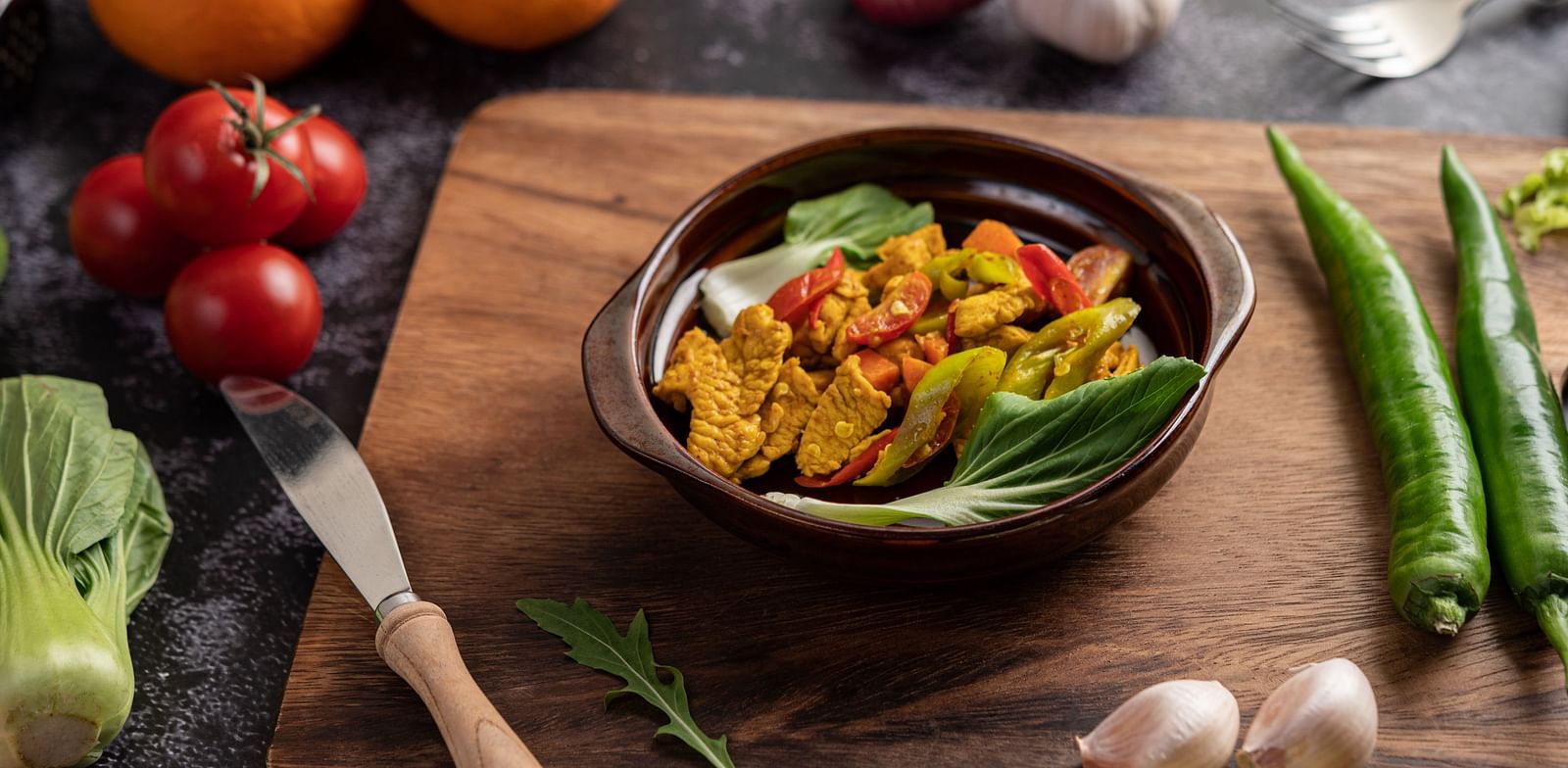 A bowl of cooked food is placed on a wooden board amidst fresh vegetables.