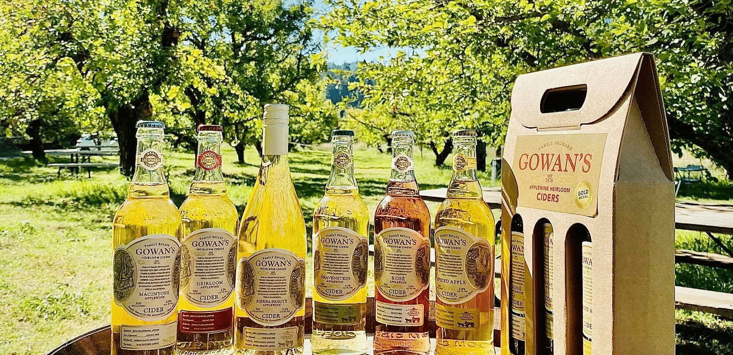 Bottles of Gowan's cider and a gift box sit on a barrel in an orchard with fallen leaves and a bright sky.
