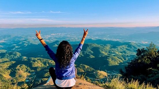 a girl sitting on top of a hill looking at the nature below and the blue sky in the background