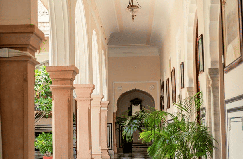 A sunlit corridor with monochrome flooring, arched walls, and potted plants along the walkway - Khas Bagh, Jaipur.